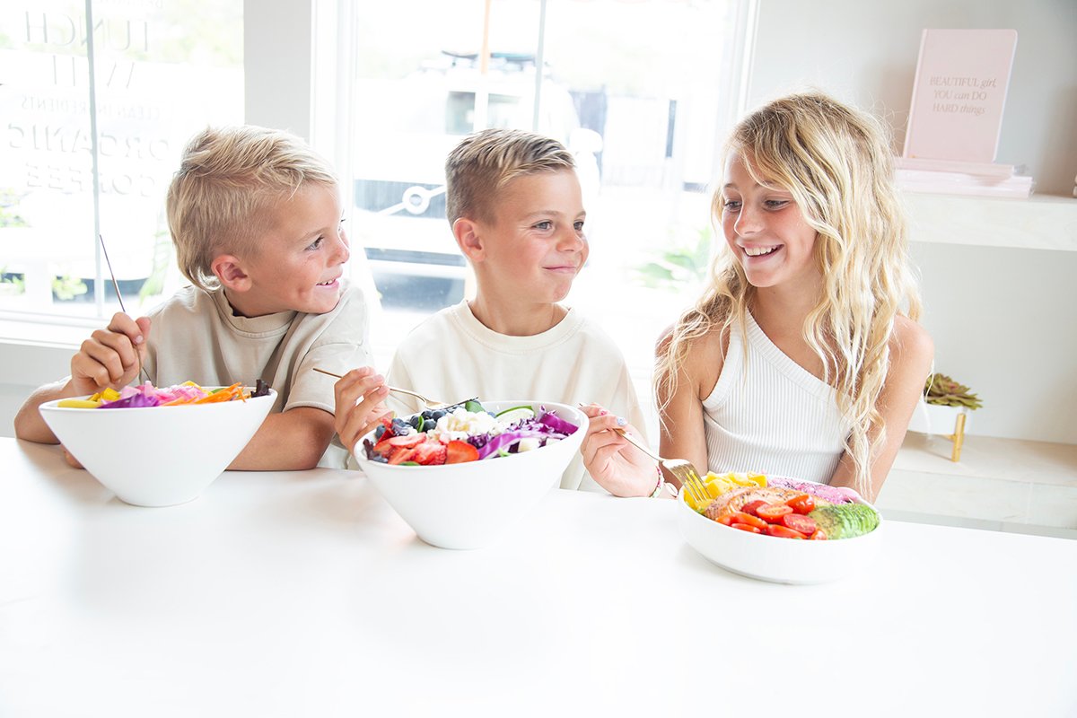 three siblings sitting side by side at white table wait to eat bowls of fruit salads