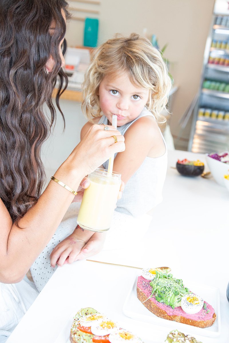 blonde hair girl looks with eyes to camera while mom offers her straw for smoothie
