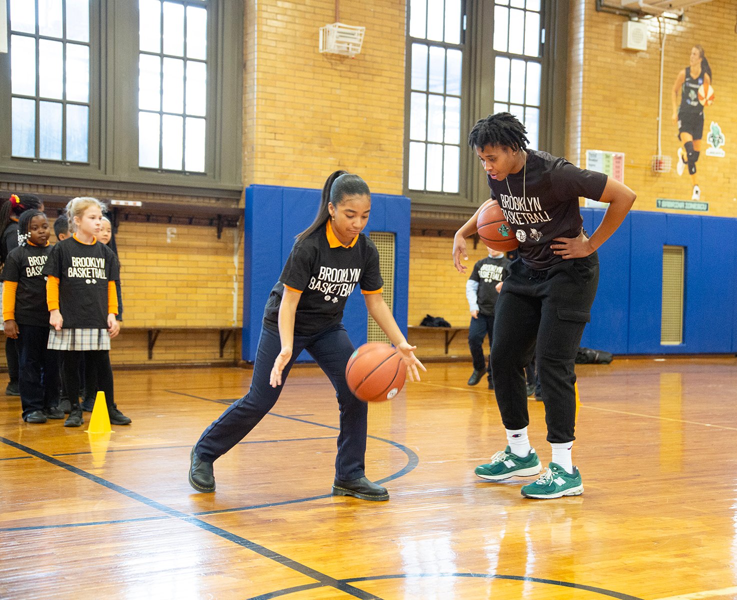 girl shows how she dribbles basketball for teacher with New York Liberty decal on gym wall