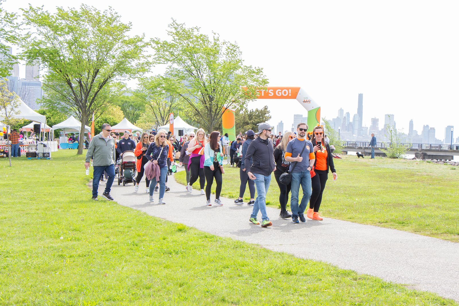 hundreds of people including the event's spokeswoman in an orange shirt walk past white tents and lower Manhattan skyline