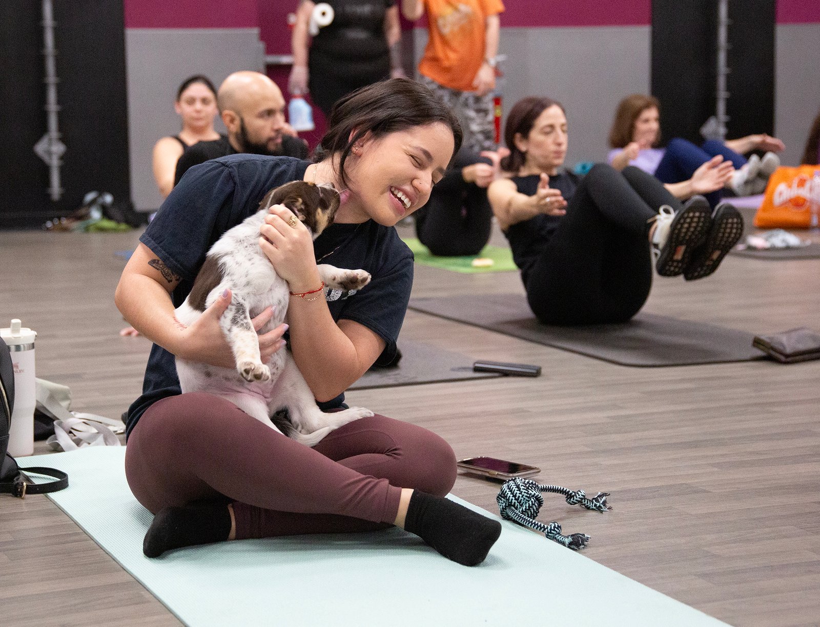 puppy licks woman sitting on mat surrounded by Stanley cup and dog toys, not striking same pose as women in background