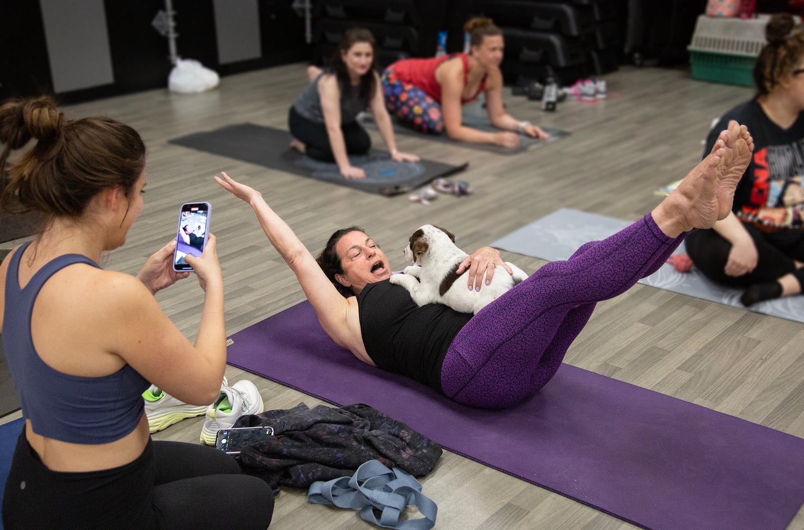 woman holds feet and arms off ground in modified boat pose with puppy on her stomach while friend takes picture