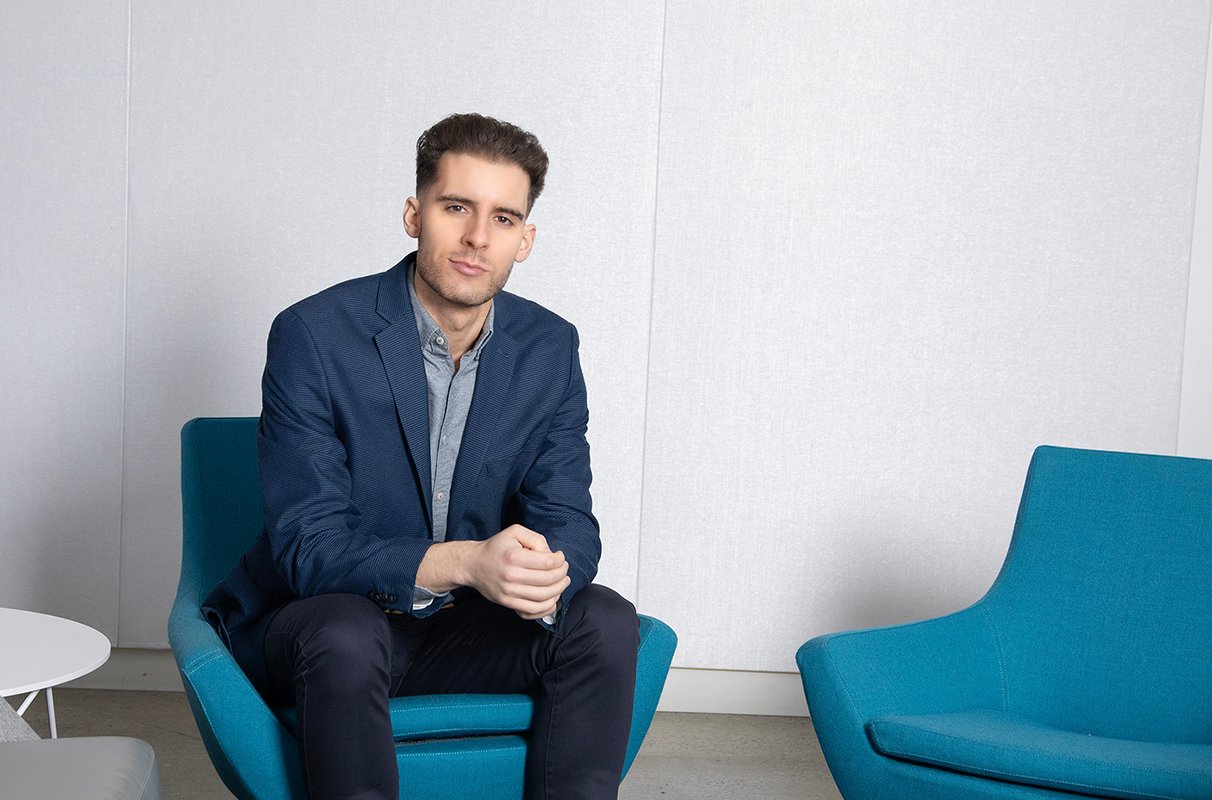 man sits in blue chair with empty blue chair next to him, smiles at camera